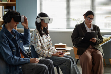 Three teenagers sitting in counseling session, two multiethnic teenagers wearing virtual reality headsets while female counselor with clipboard observing and taking notes nearby