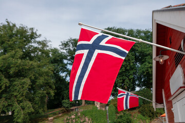Two Norwegian flags hang from poles on a red wooden building, fluttering in breeze against a...