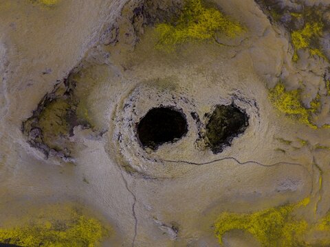 Aerial view of dark voids puncturing the pale, cracked earth, accented by patches of vibrant yellow moss or lichen, creating a surreal, otherworldly landscape., Location Omitted, Location Omitted.