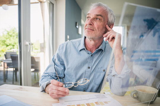 Thoughtful senior freelancer with transparent screen sitting at table