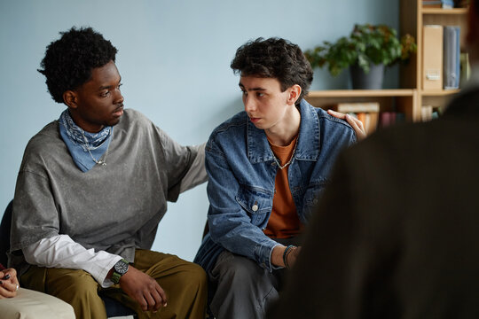 Multiethnic group of teenage boys sitting together during counseling session, Black teenager comforting Caucasian teenager by placing hand on shoulder, both showing serious expressions