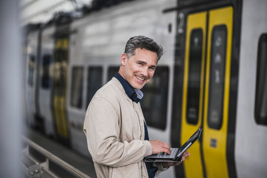 Happy businessman with tablet PC standing in front of train at railroad station
