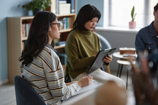 Multiethnic teenage girls participating in counseling session, sitting in chairs, writing on clipboards, engaging in group therapy activity with another partially visible - Powered by Adobe