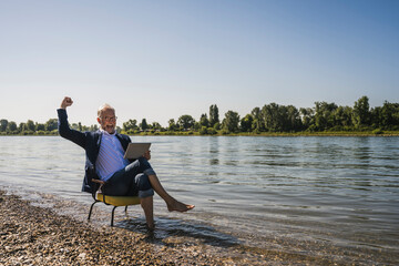 Happy senior man holding tablet PC sitting on chair gesturing at riverbank