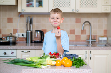 Happy boy holding strawberry near fruits and vegetables in kitchen. Natural snack and healthy baby food concept