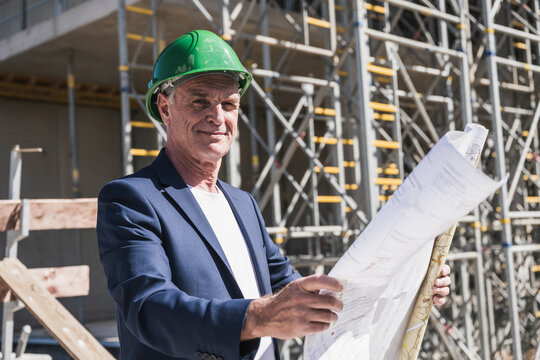 Smiling architect with blueprint standing at construction site