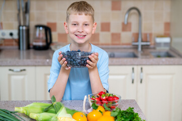 Blond teenage boy holding bowl of blueberries in bright kitchen with fresh fruits