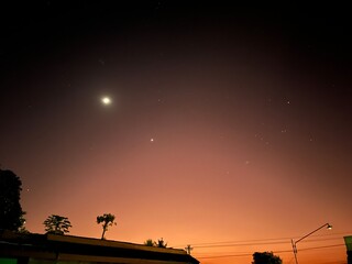 Night sky with crescent moon and bright planet above rooftops during twilight, featuring stars and a warm orange horizon.