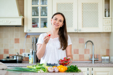 Young woman holding bowl of strawberries in bright kitchen with fresh vegetables and berries