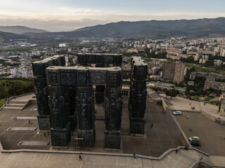 Aerial view of the Chronicles of Georgia's dark, monolithic pillars under a soft sky, contrasting with the distant city, Tbilisi, Tbilisi, Georgia.