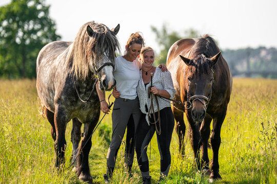Two joyful young women spending time with their horses in lush green field on sunny day. Cheerful and heartwarming rural scene that captures friendship, emotional bond between people and animals. - Powered by Adobe
