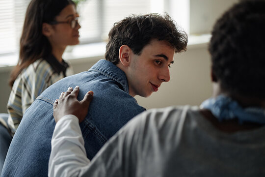 Caucasian teenage boy sitting in counseling session with diverse group of teenagers, Black teenage boy offering support by placing hand on his shoulder, another teenage girl in background listening