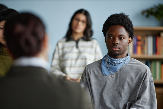 Black teenage boy sitting in counseling session with multiethnic group of teenagers listening attentively to adult woman, bookshelf in background, focus on mental health support - Powered by Adobe