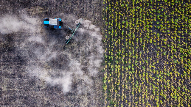 Aerial view of a lone blue tractor kicking up dust in a freshly plowed field next to a crop field, Mykolaiv, Mykolaiv Oblast, Ukraine.