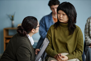 Asian teenage girl sitting with hands clasped while female counselor holding clipboard engaging in...