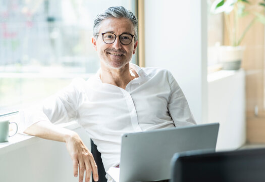 Smiling businessman sitting with laptop leaning head on window sill at office