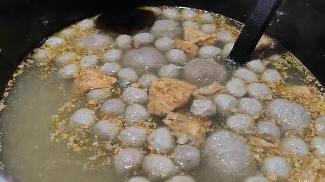 A close-up video of traditional Indonesian meatballs (bakso) inside a large metal steamer at a street food stall. The vendor prepares to serve the hot meatballs into bowls for customers. This scene ca