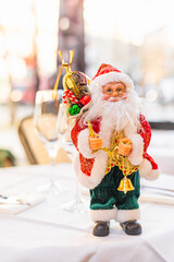 A decorative Santa Claus figurine stands on a table. The Santa has a white beard, red suit, and holds bells and a sack. The background is softly blurred.