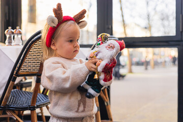 A young Caucasian girl with blonde hair wears a reindeer headband. She holds a Santa Claus toy in a festive setting, celebrating Christmas and New Year.