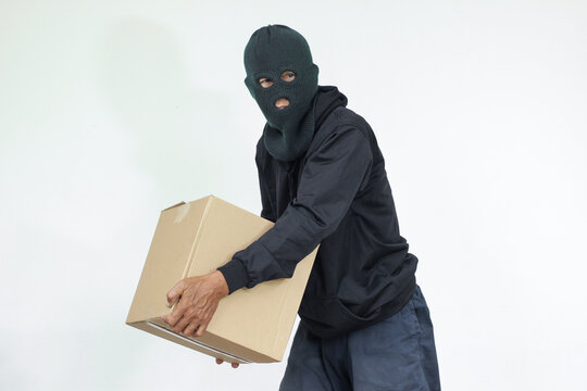 A masked figure steals a cardboard box in a house. The thief takes valuables inside the box. The concept of package theft, shipping crime, and data thef.