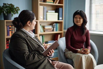 Young woman holding notebook engaging in conversation with Asian teenager sitting in counseling session, both women facing each other in professional setting