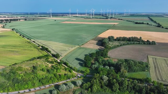 Aerial view of vibrant patchwork fields contrasting with dark green trees and distant wind turbines under a clear blue sky, Brussow, Brandenburg, Germany.