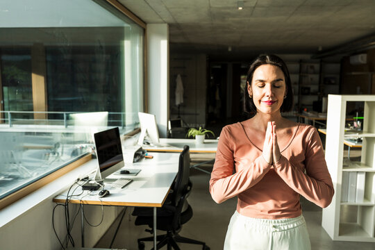 Smiling mature businesswoman with eyes closed and prayer pose standing in office