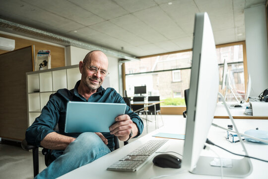 Mature businessman using tablet PC sitting at computer desk in office