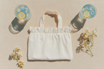 A white cotton tote bag with soft ruffled edges rests on a beige linen tablecloth. Surrounding it are light summer flowers and refined cocktail glasses filled with ice cubes and lemon slices