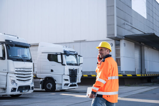 Smiling mature dock worker wearing protective workwear talking on mobile phone at warehouse