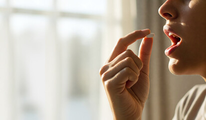 Soft morning light illuminates a woman's hand gently guiding a pill to her mouth, a close-up of health and self-care