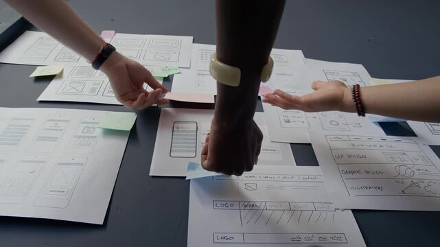 Close up on hands of diverse creative team discussing UI design of mobile app pointing at paper wireframes handing on wall while collaborating in office space