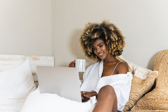 Happy young multiracial latina woman sitting on bed typing on laptop while having morning coffee - Powered by Adobe