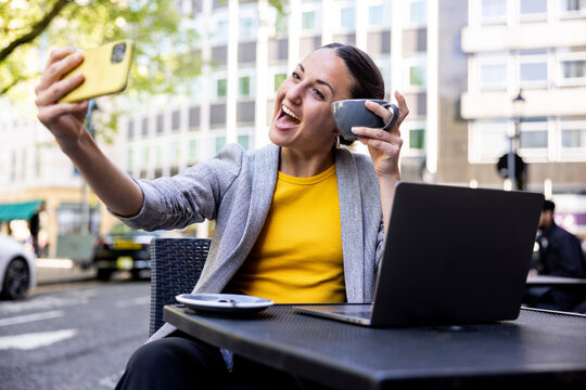 Happy businesswoman holding coffee cup taking selfie through smart phone at sidewalk cafe