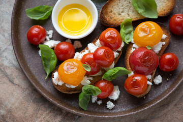Bruschettas with roasted mixed tomatoes, ricotta cheese and green basil served on a brown plate,...