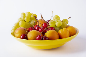 Cherries, apricots and grapes lie on a yellow ceramic plate on a white background