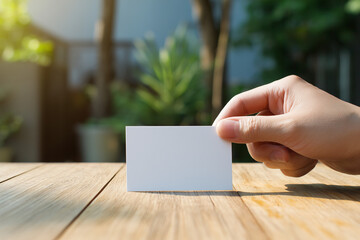 Hand holding a blank white card on a wooden table outdoors with blurred garden background.