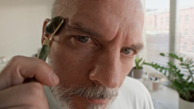 Bearded senior man looking in mirror while standing in spacious daylit bathroom and making massage of face with jade roller during self care ritual in the morning - Powered by Adobe