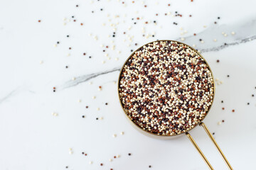 Tri-color quinoa seeds in a measuring cup on white kitchen table. Top view. Copy space.