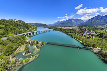 Aerial view of turquoise waters embracing bridges connecting verdant banks under a vast blue sky, Drava River, Ferlach, Carinthia, Austria. Beautiful landscape view on a picturesque summer day.