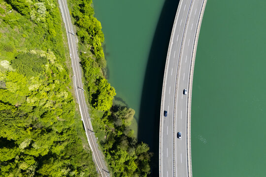 Aerial view of a bridge curving gracefully over the vivid turquoise Drava River, contrasting with the lush green forest and parallel railway line, Ferlach, Carinthia, Austria.