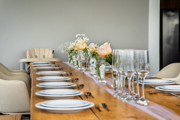Long wooden dining table set with white plates, silver cutlery, glassware, and soft peach rose centerpieces in a bright, modern indoor setting.