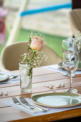Romantic table setting with single blush rose and baby's breath in glass vase with white stones, soft natural tones, green plate, glassware, minimal wedding or outdoor event ambiance.