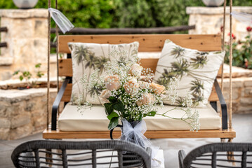 Floral bouquet with blush roses and baby's breath on a rustic wooden bench with tropical-patterned pillows in a peaceful outdoor garden.