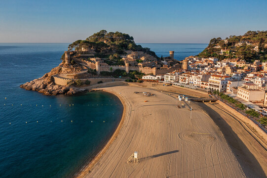 Aerial view of the golden beach curving towards the medieval castle walls, bathed in the warm glow of the morning sun, Tossa de Mar, Catalunya, Spain.