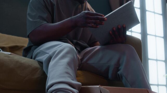Tilt up shot of young Black man with electronic monitoring ankle bracelet sitting on couch and looking through photo album, reminiscing about past under house arrest