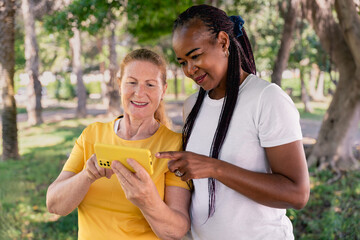 Two joyful multi-ethnic women sharing moments while using a smartphone in a park, celebrating technology and the bond of friendship