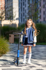 Little girl stands with scooter in front of multi-storey building background. School girl riding a scooter in city. Active leisure and outdoor sport for child. Eco-friendly urban transport for kid.
