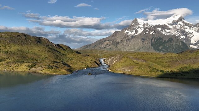 Aerial view of glacial meltwater cascading through rugged, green hills into the serene blue lake beneath snow-capped mountain peaks, Torres del Paine, Magallanes, Chile.