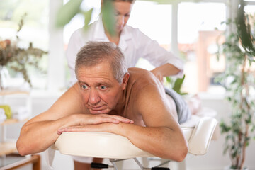 Glad elderly man having back massage procedure done by middle-aged masseuse in therapy cabinet